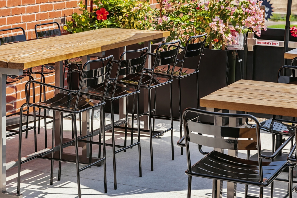 Black barstools up against a table with a wood top and silver steel legs, next to it is a matching dining-height table and chairs
