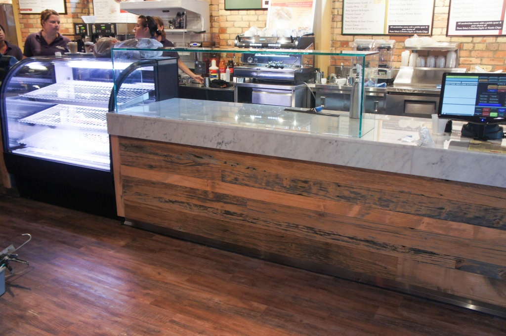A servery system with a wooden dinoc finish on the base, grey corian top, and glass sneezegaurd.