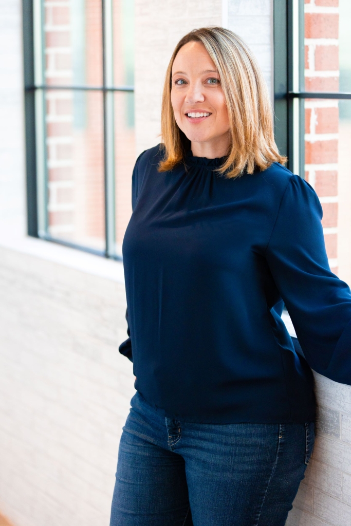 Andrea Hubbard smiling and wearing a blue shirt leans up against a window