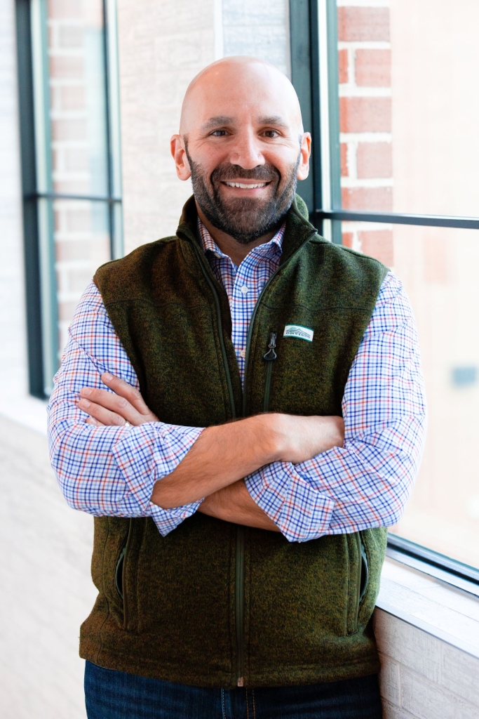 Casey Cain smiling with his arms crossed wearing a blue and red checkered shirt with a grey zip-up vest