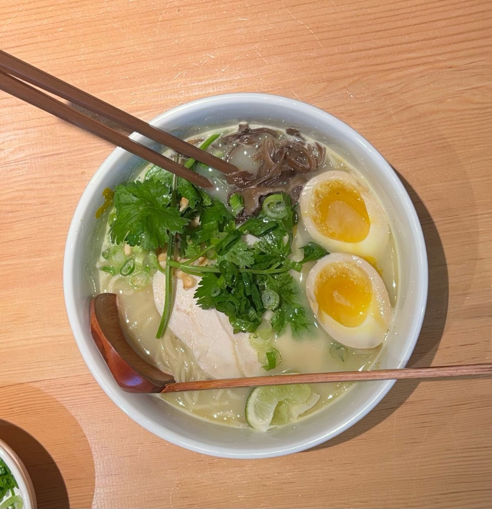 A white bowl holds a serving of coconut curry ramen with mushrooms, a seasoned egg, chicken, scallions, a lime wedge, chopsticks and a wooden spoon.