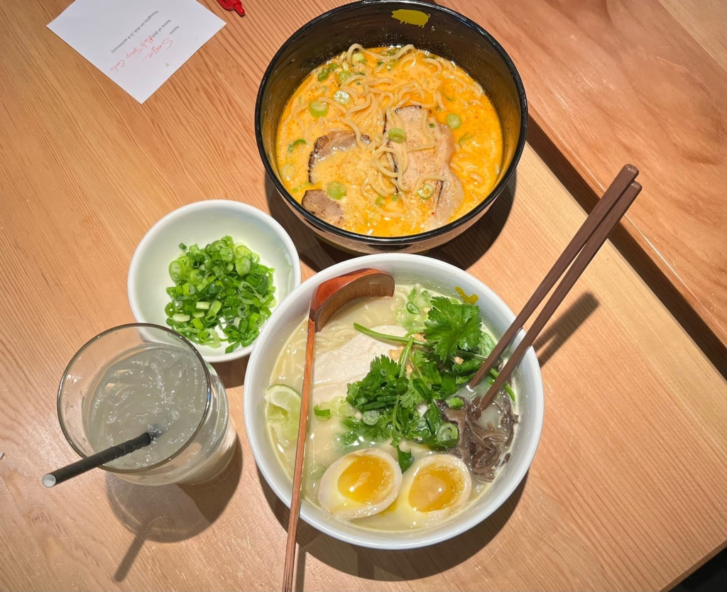 Two bowls of ramen, a small bowl of scallions, and a glass of lemonade with a black straw.