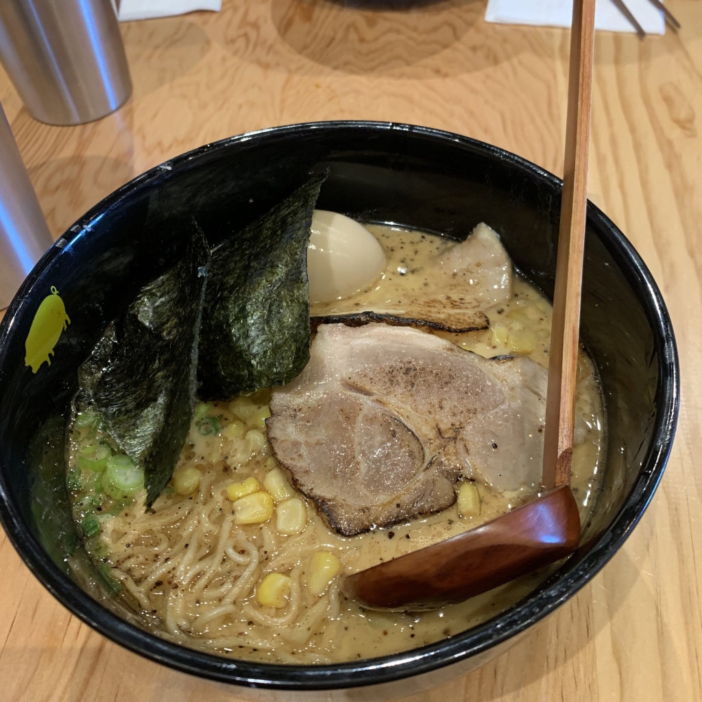 A black bowl holds a serving of Pork Miso Ramen, with noodles, corn, nori sheets, scallions, and a wooden spoon.
